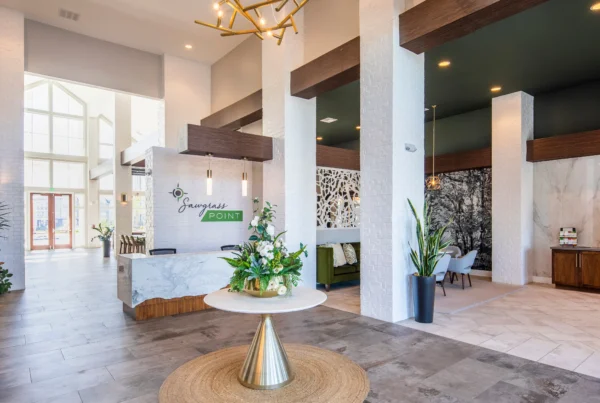 The image depicts the elegant interior of the Sawgrass Point lobby. It features a high ceiling, modern decor with white walls, and a central table adorned with a floral arrangement. The space includes a reception desk with the Sawgrass Point logo, contemporary furnishings, and a coffee station, creating a welcoming and luxurious atmosphere.