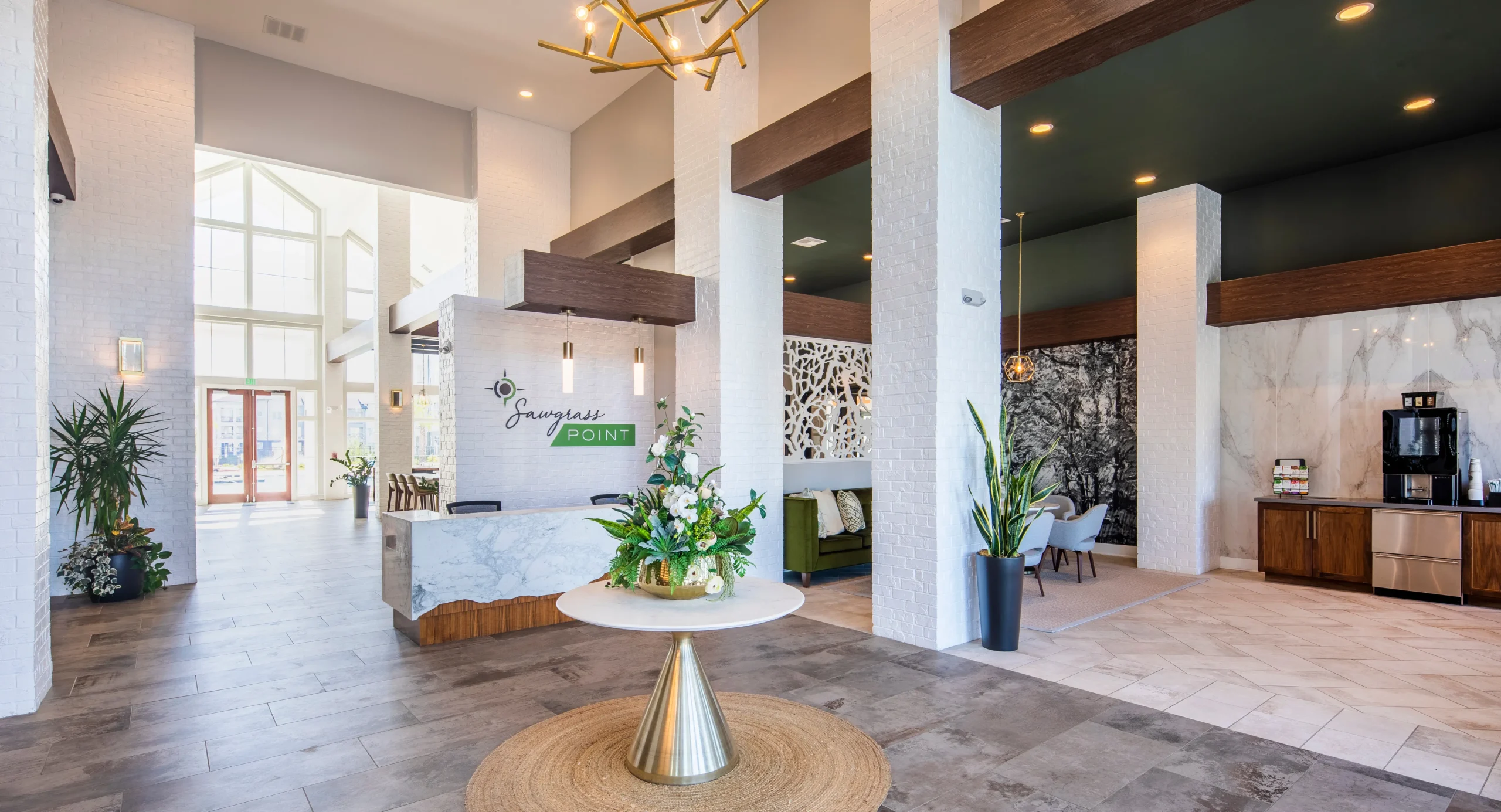 The image depicts the elegant interior of the Sawgrass Point lobby. It features a high ceiling, modern decor with white walls, and a central table adorned with a floral arrangement. The space includes a reception desk with the Sawgrass Point logo, contemporary furnishings, and a coffee station, creating a welcoming and luxurious atmosphere.